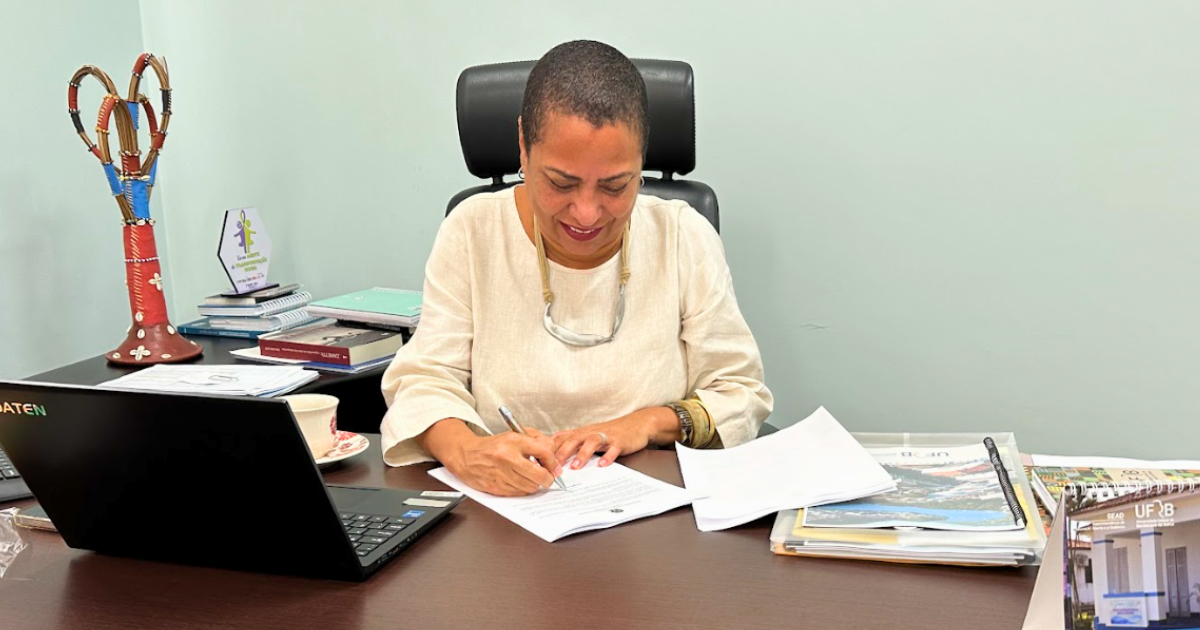 A imagem mostra uma mulher sentada em uma mesa de escritório, assinando documentos. Ela tem pele morena, cabelo curto e veste uma blusa clara. A mesa está cheia de papéis, livros e um laptop preto à sua esquerda. Há também uma xícara de café com pires ao lado do laptop. No fundo, à esquerda, há um objeto decorativo colorido e alguns livros empilhados. A mulher parece concentrada no que está fazendo e está sorrindo levemente.